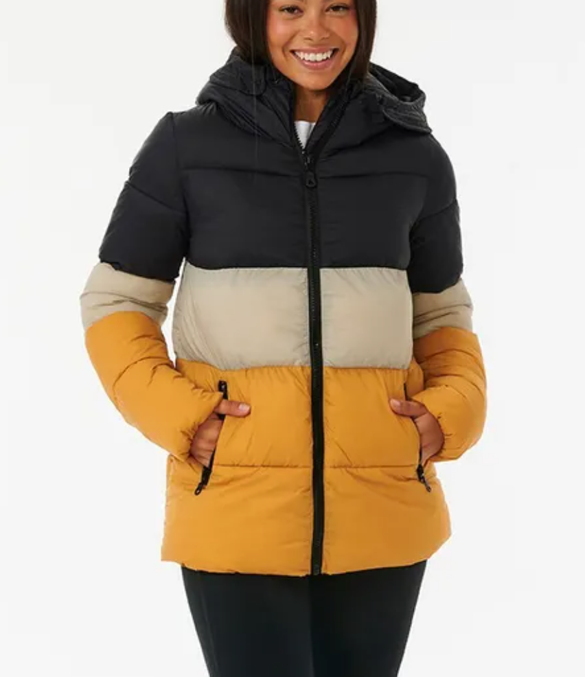 A woman smiles in a Rip Curl Anti Series Insulated II Jacket, showcasing a color-blocked design with black, beige, and mustard yellow horizontal sections. With a hood and water-repellent technology, she stands confidently with her hands in the front pockets.