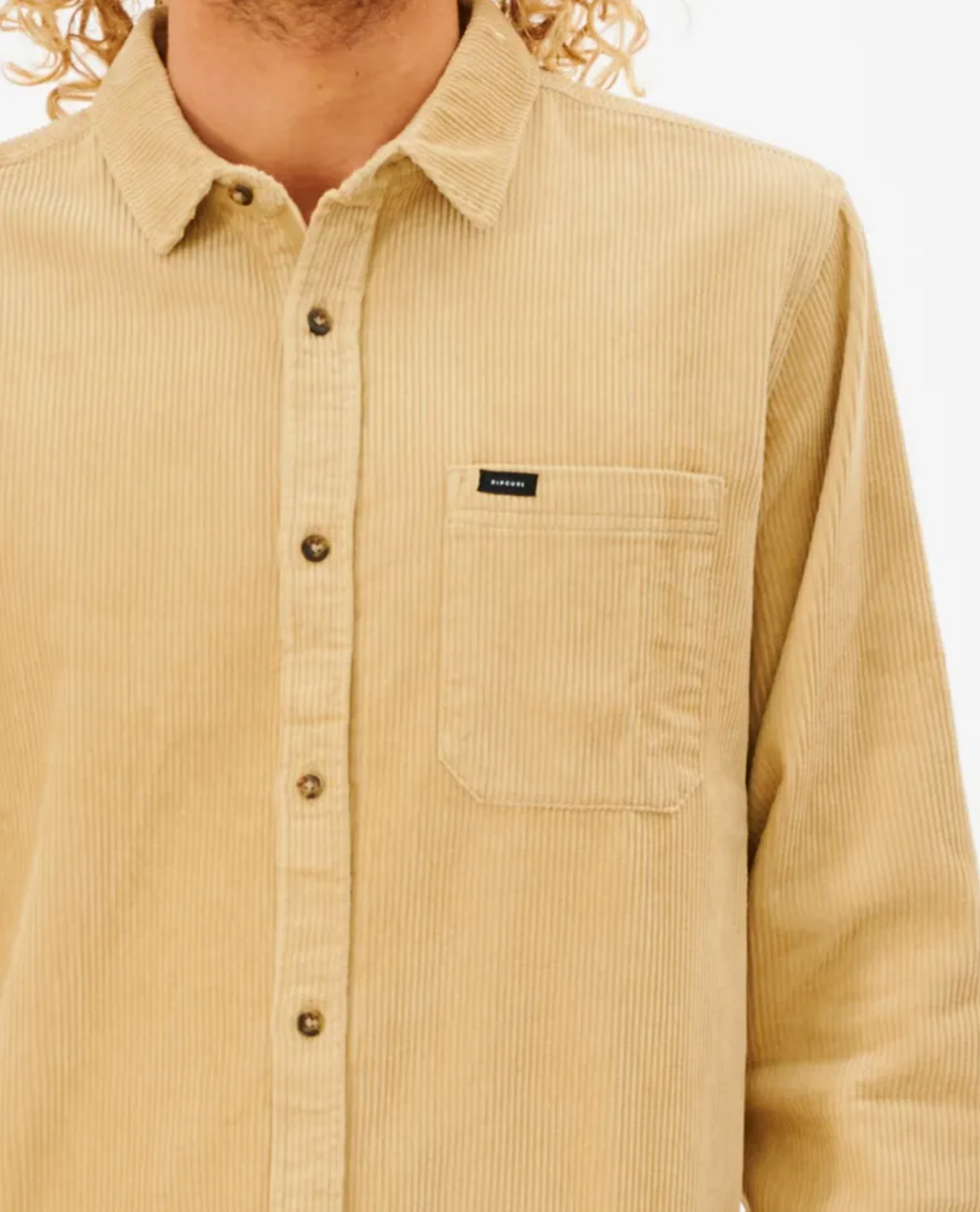 A person in a Rip Curl State Cord Shirt, a beige cotton corduroy long-sleeve with a front pocket and dark buttons, stands against a white background, flaunting their curly hair.