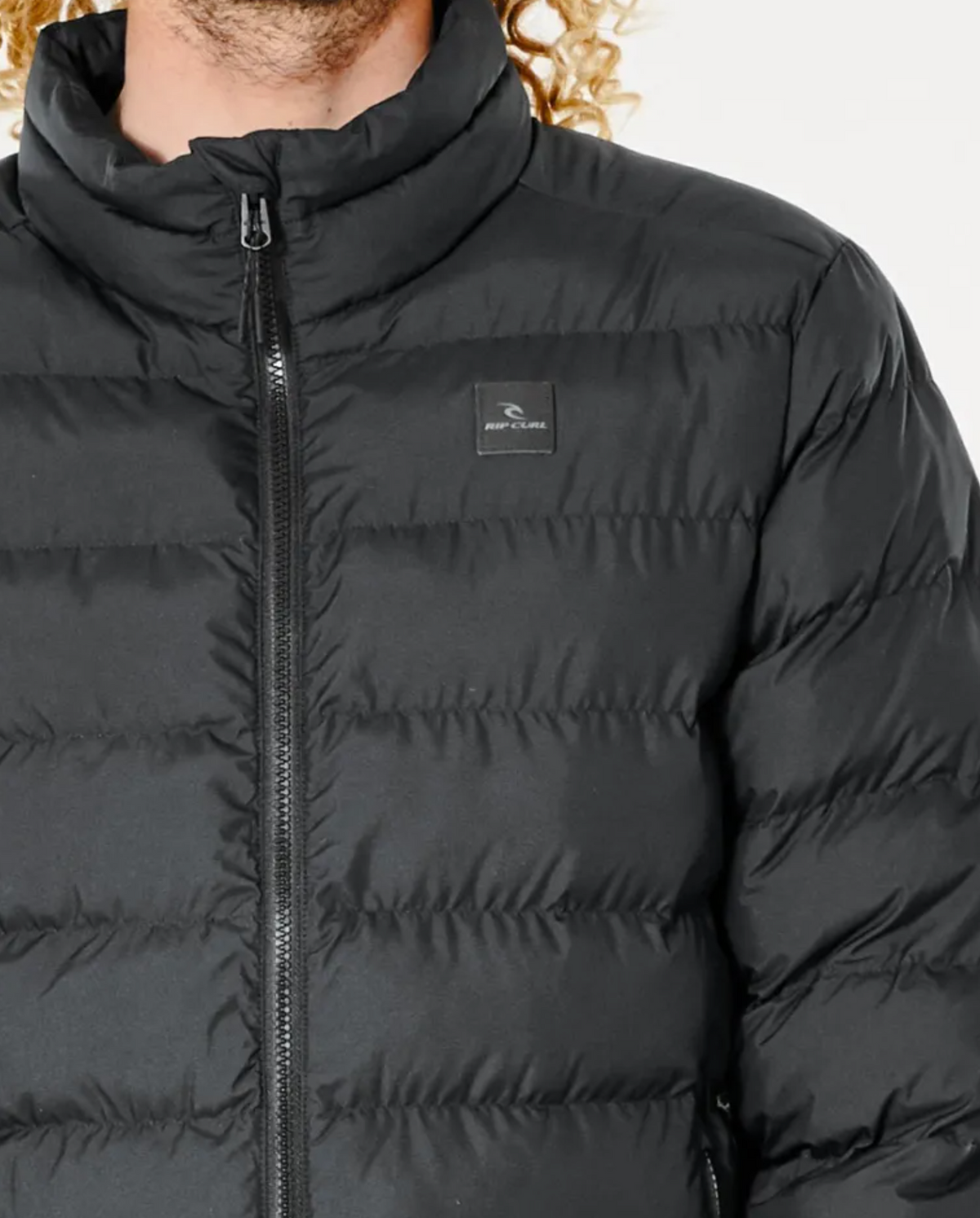A person is wearing a Rip Curl Anti Series Elite Puff Crew Jacket, made from premium duck down, featuring a black puffer design with a high collar and visible zipper. A small logo with a bird silhouette adorns the chest. Curly hair peeks out, adding to the stylish look.