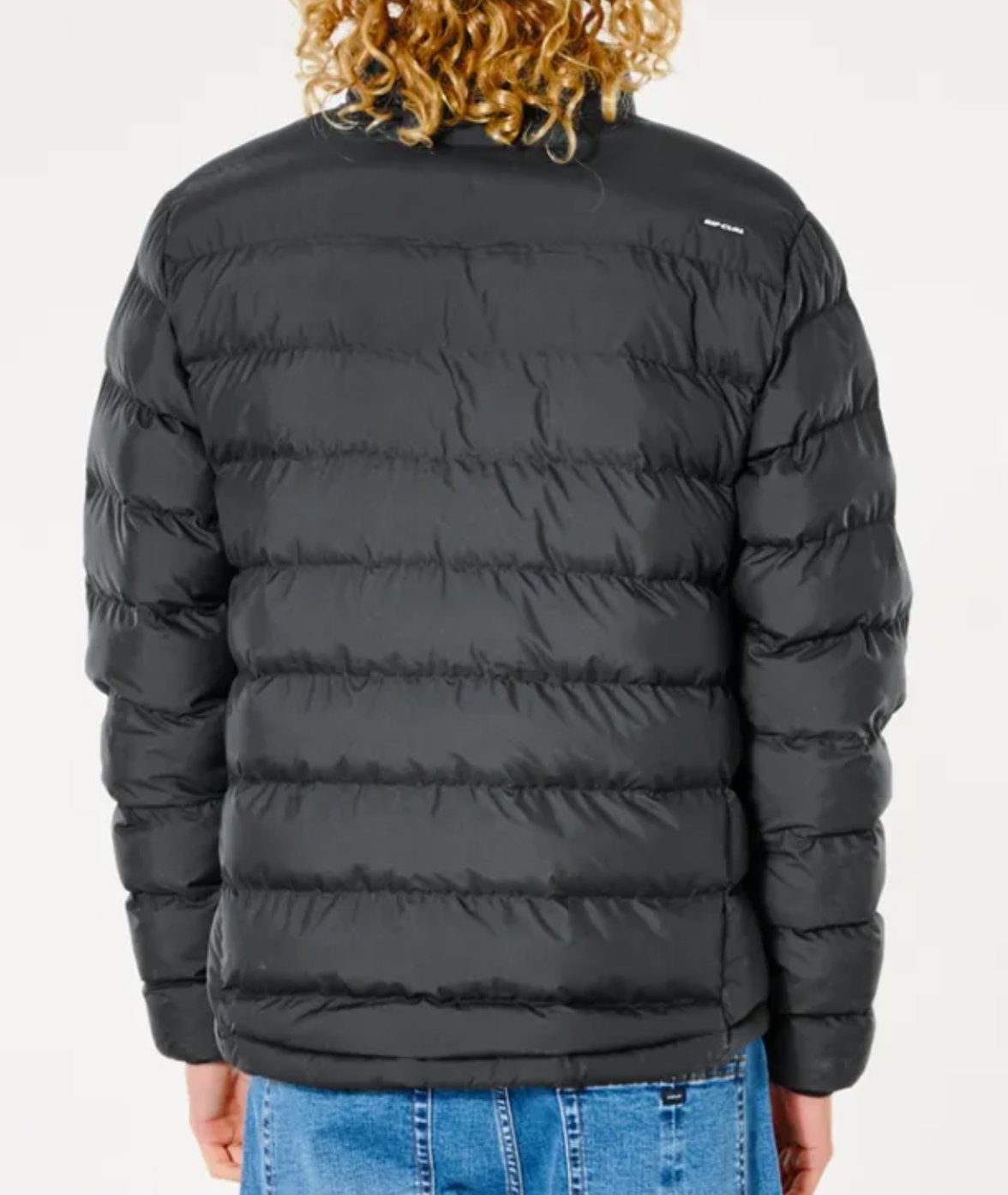 A person with curly hair is wearing a Rip Curl Anti Series Elite Puff Crew Jacket in black, filled with premium duck down, and blue jeans, facing away from the camera.