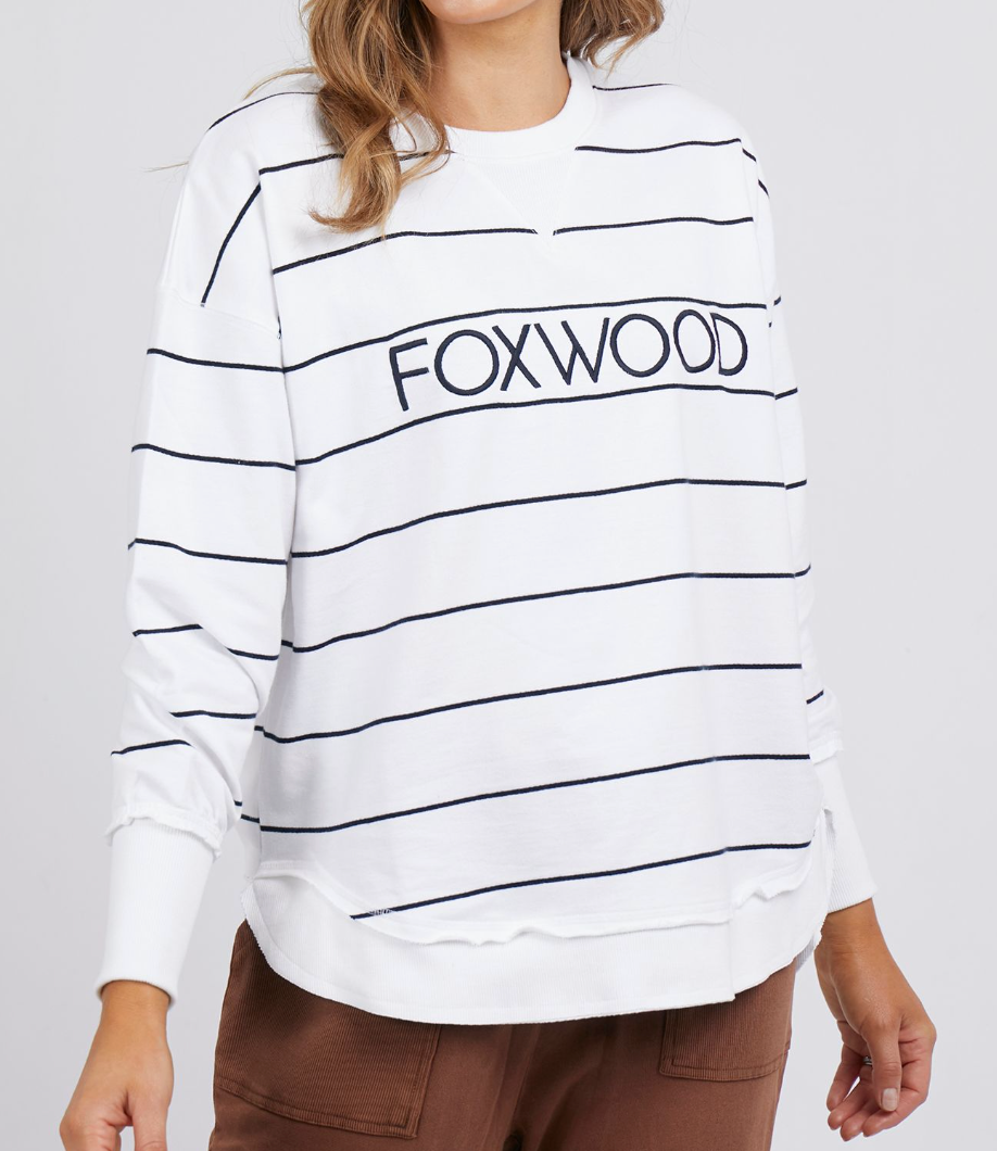 The person wears the Foxwood Simplified Stripe Crew—a relaxed fit white sweatshirt with black stripes and "FOXWOOD" lettering, crafted from 100% cotton. It's paired with brown pants for a stylishly comfortable look.
