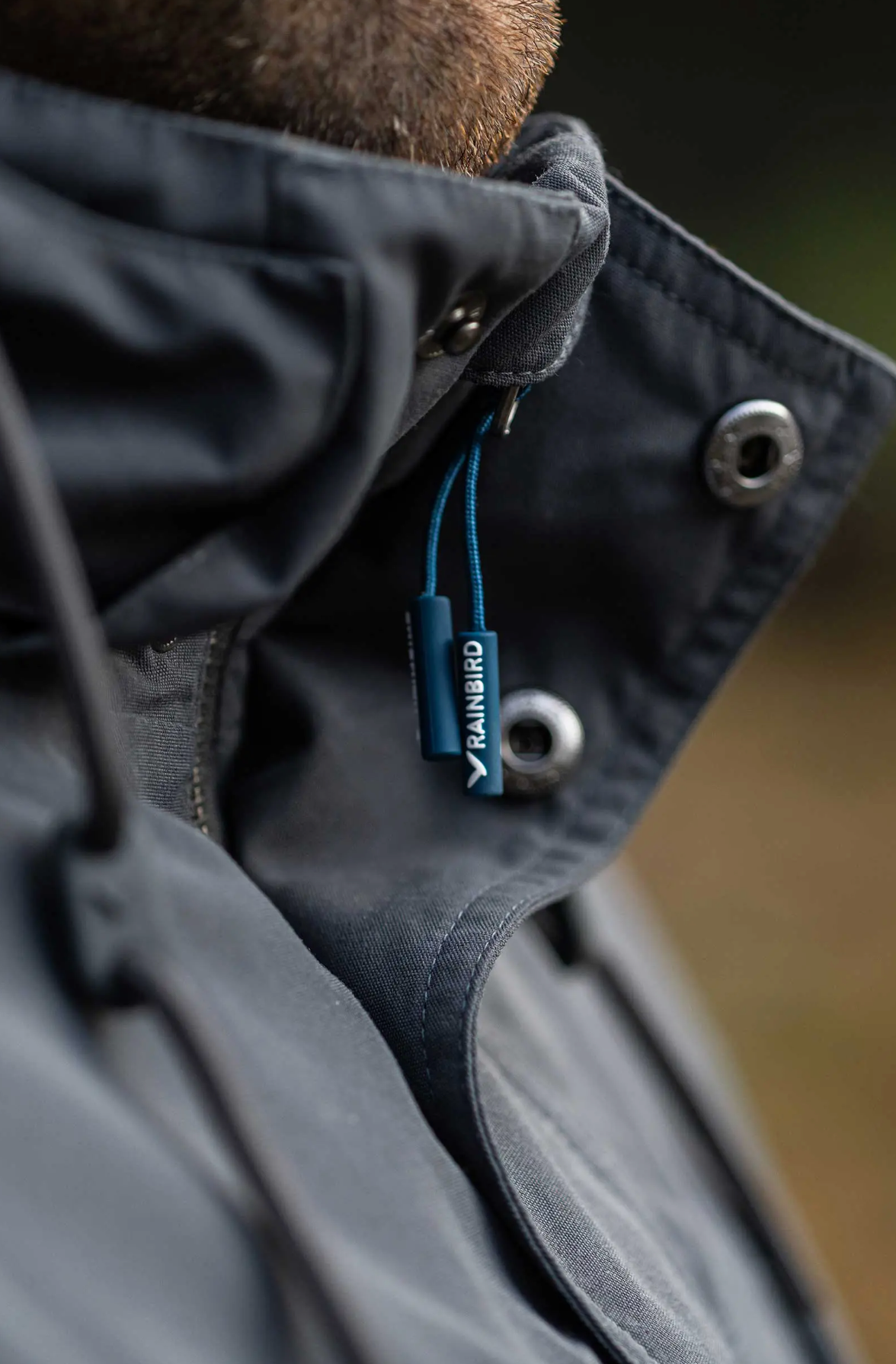 Close-up of a person wearing the windproof Rainbird Altair Jacket, featuring a high collar and dark waterproof fabric. A branded zip pull labeled "RAINSIRD" is partially visible, secured to a blue drawstring, highlighting its quality craftsmanship against a blurred background.