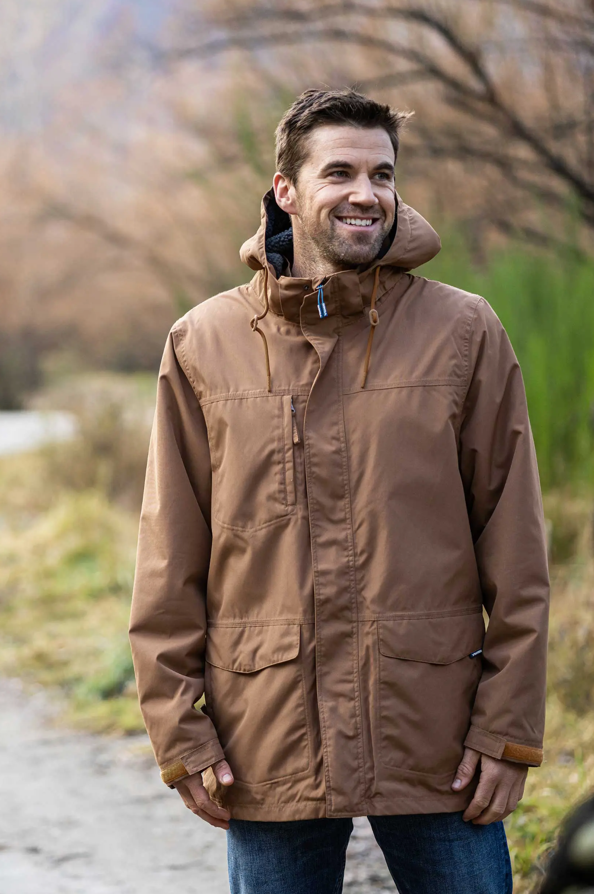 A person outdoors on a pathway wears a brown Rainbird Altair Jacket with fleece lining. Blurred trees and foliage provide a natural backdrop as they smile and gaze slightly to the side.
