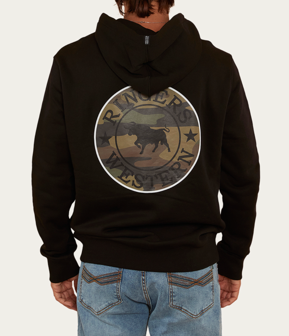 The person wears the Ringers Western Signature Bull Hoodie, a black cotton piece with a circular bull emblem and "Ringers Western" in camouflage on the back. Paired with blue jeans, the ensemble offers a casual yet stylish look.