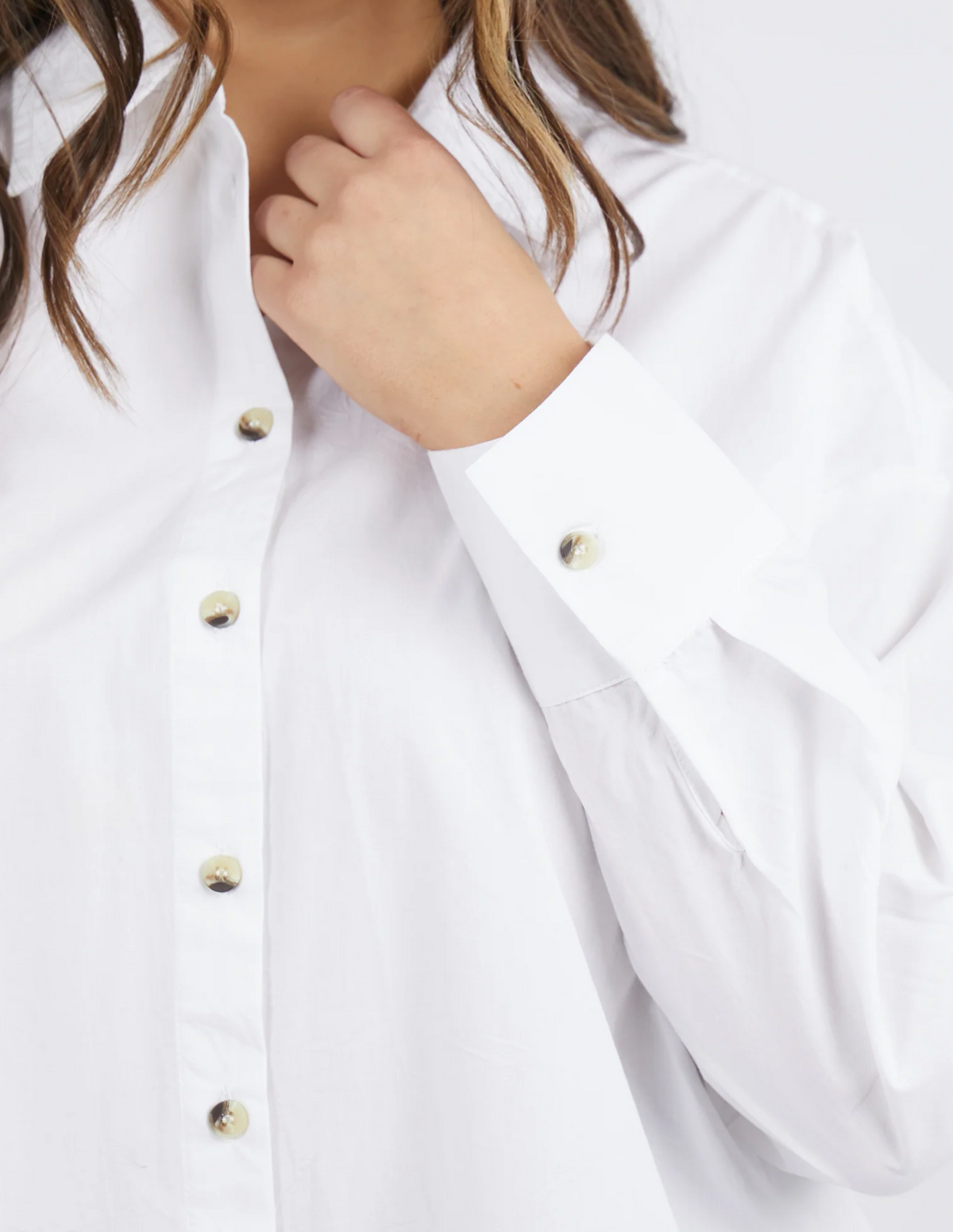 A person wearing a stylish Foxwood Paulina Shirt, made from 100% cotton poplin, adjusts the collar with their left hand. This white button-up design features gold buttons and long sleeves, set against a plain white background.