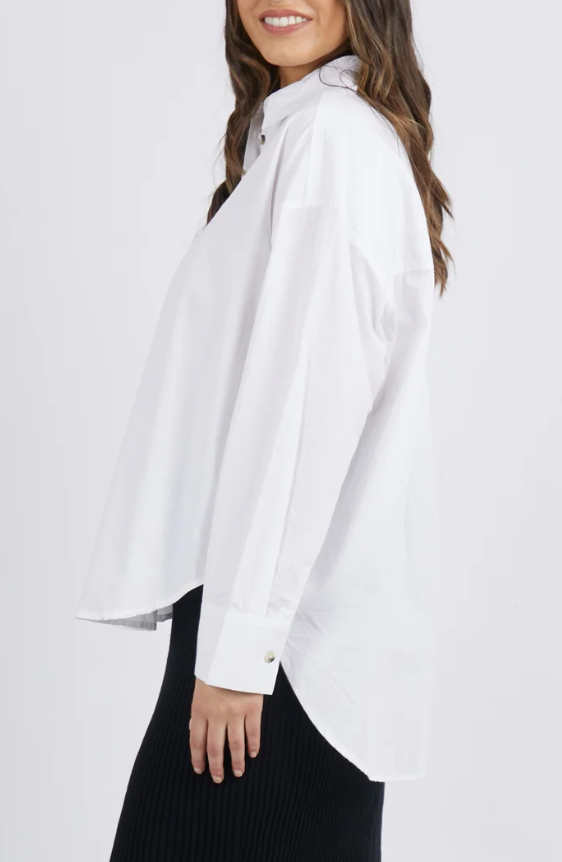Side view of someone in a Foxwood Paulina Shirt, a 100% cotton poplin button-up with long sleeves, paired with a black skirt. They have long, wavy brown hair and are smiling against a crisp white background.