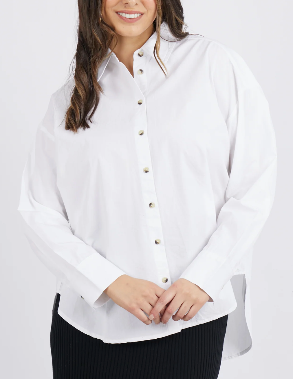 A person wearing a stylish Foxwood Paulina Shirt, made from 100% cotton poplin, and dark pants stands against a white background. The slightly oversized shirt has long sleeves and buttons down the front. The individual with long, wavy brown hair is smiling.