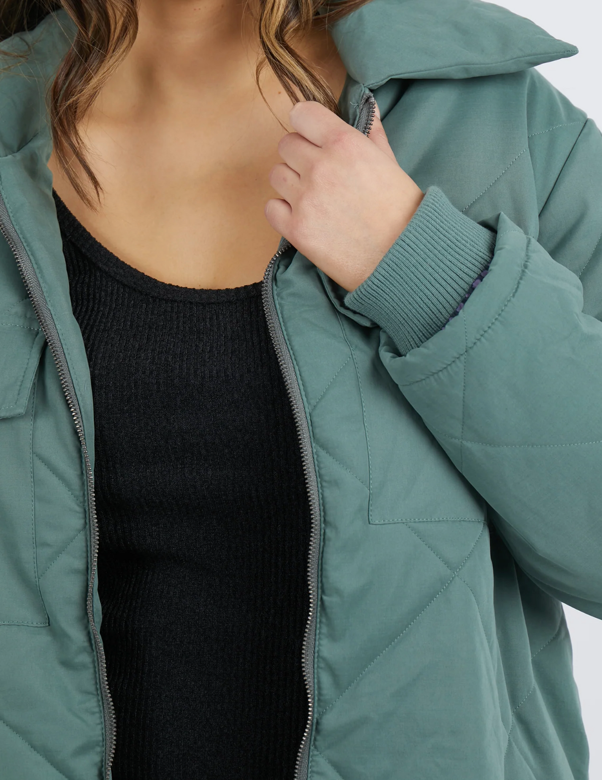 A person with wavy hair is partially visible, wearing the teal Foxwood Whitley Jacket over a black ribbed top. They hold the collar, slightly pulling it closed to reveal subtle design elements like zippered side splits for flair and function.