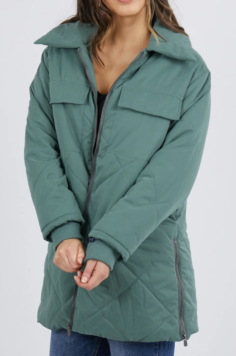 A person wears a teal-quilted Foxwood Whitley Jacket with a front zipper, large collar, and two chest pockets, hands together and face partially visible. This slightly oversized jacket features zippered side splits, paired stylishly with blue jeans.