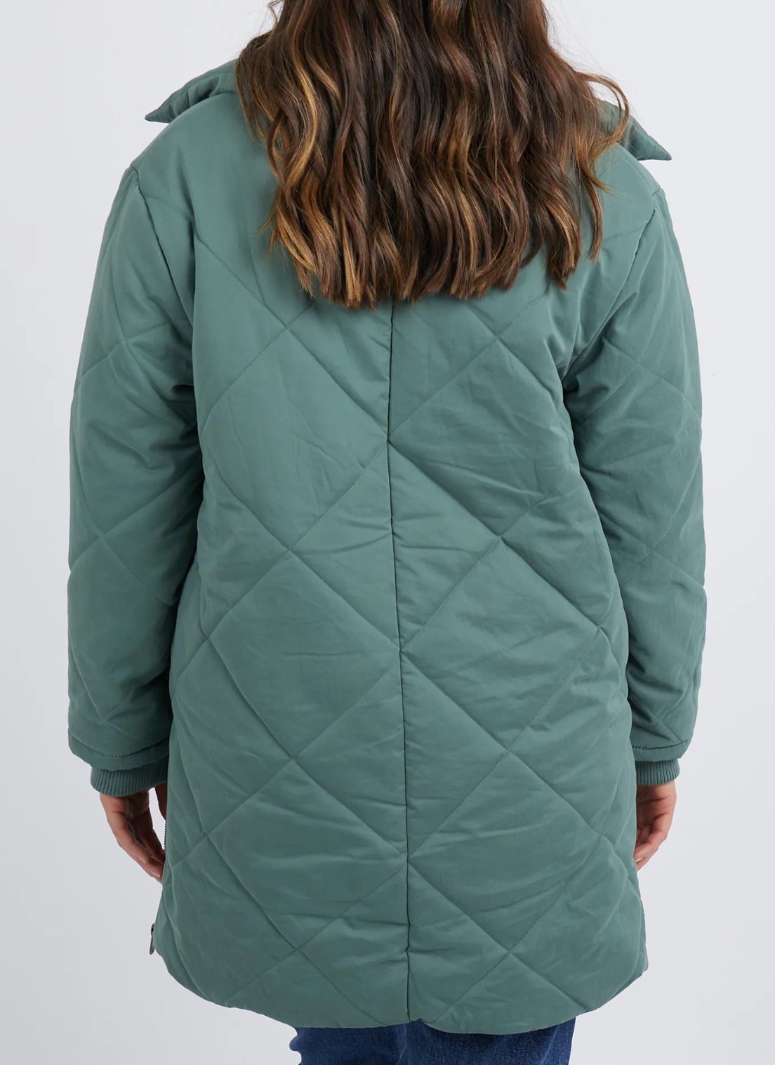 A person with wavy brown hair faces away from the camera, wearing a cozy Foxwood Whitley Jacket — a quilted green coat featuring a collar and zippered side splits. Paired with blue jeans, they stand against a plain, light-colored background.
