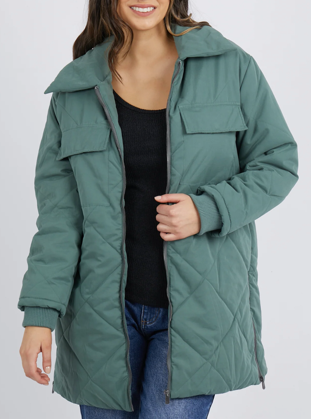 Against a plain backdrop, a person in a cozy teal quilted Foxwood Whitley jacket and black shirt displays its stylish design with one hand. The jacket includes zippers, two chest pockets, and zippered side splits.