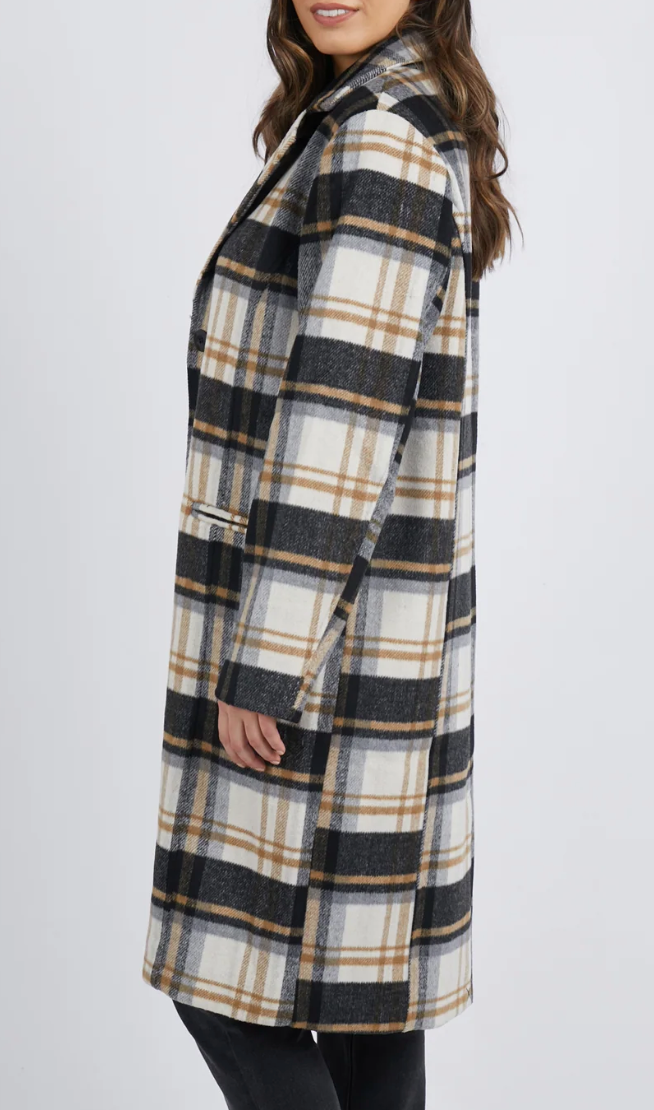 Wearing a Foxwood Catriona Coat in a chic plaid with black, white, and tan stripes over black pants, the person with loose wavy brown hair faces slightly sideways against a plain white backdrop. The coat’s collared design is crafted from polyester wool.
