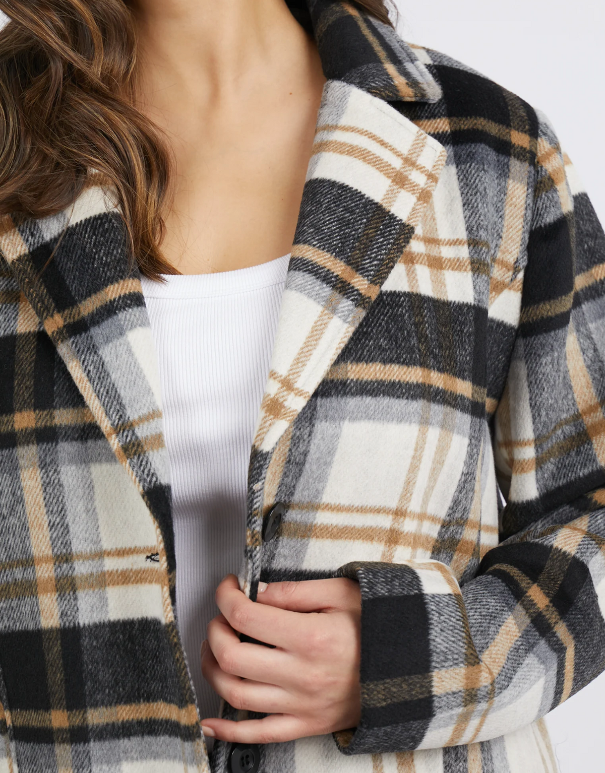 The individual adjusts the Foxwood Catriona Coat, a stylish black, brown, and white plaid piece made from polyester wool. Over a white ribbed top, they highlight its texture and pattern with their hand as their long, wavy hair cascades down one side, enhancing its allure.