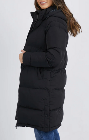 A person in side profile wears the Foxwood Camilla Puffer Jacket, a long black polyester coat with quilted detailing and a high collar. It features button details on the lower sides. The individual with long, wavy hair pairs it with blue jeans.