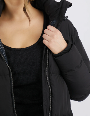 A person is shown wearing the Foxwood Camilla Puffer Jacket featuring quilted detailing, paired with a black ribbed top. The image highlights the torso, with one hand adjusting the jacket's collar and shoulder-length hair visible.