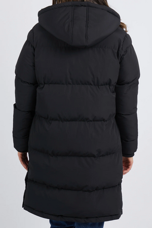 A person in a Foxwood Camilla Puffer Jacket, featuring quilted detailing and a hood, stands against a plain background. Made from polyester, this black jacket displays horizontal stitching for an insulated appearance.