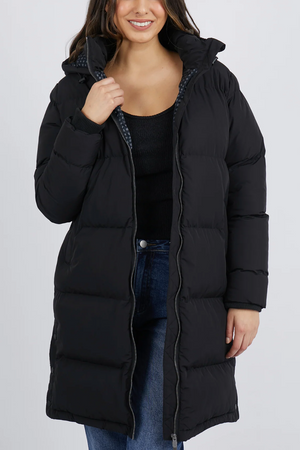 Wearing a Foxwood Camilla Puffer Jacket in long black with quilted details, a person pairs it with blue jeans and a black shirt. The polyester jacket, featuring a high collar and patterned lining, is unzipped against the plain white backdrop.