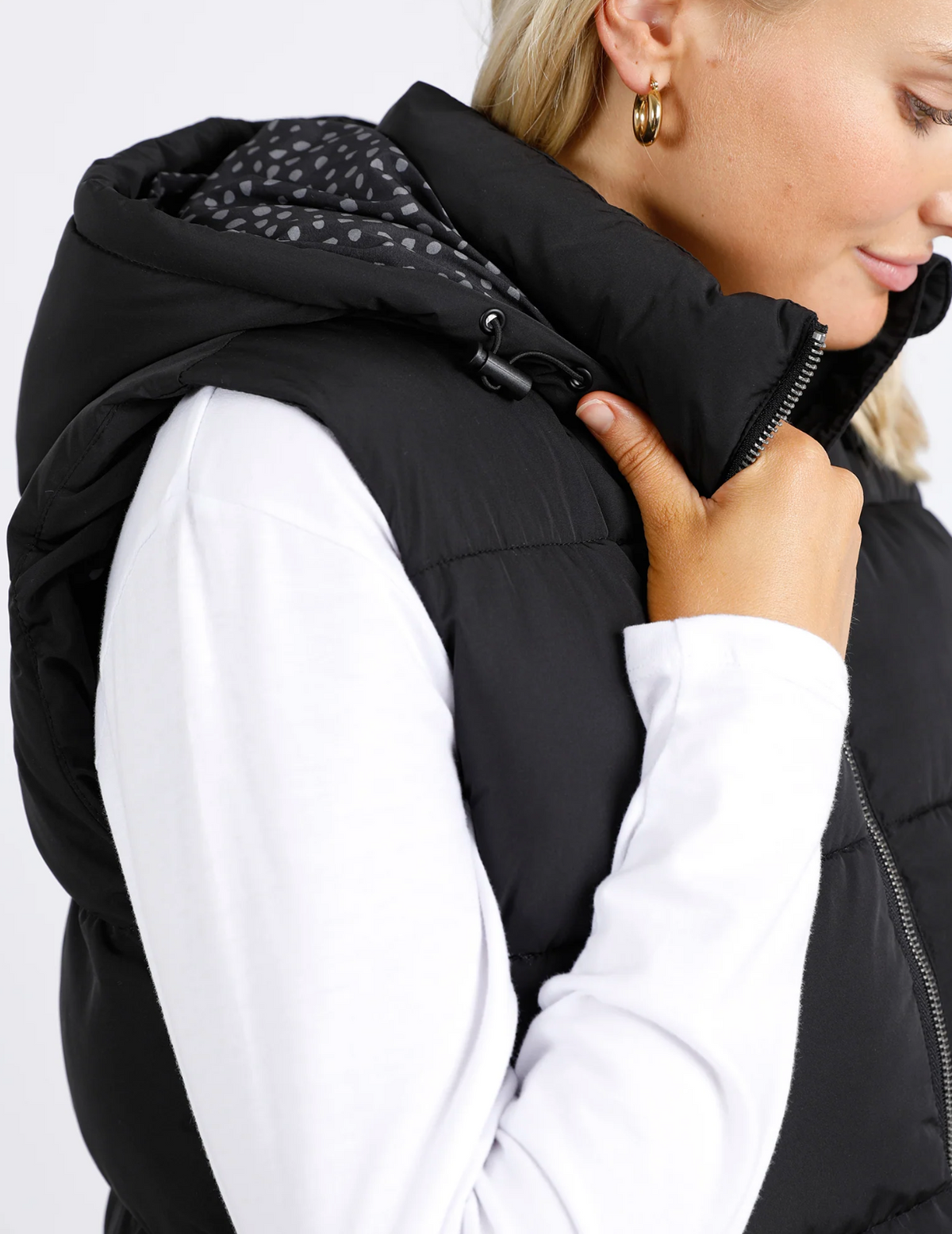 A person wearing the Foxwood Camilla Puffer Vest in black with quilted detailing and a white long-sleeve shirt. They're adjusting the cozy vest's collar, revealing a patterned lining. The look is completed with an adjustable hood and gold hoop earrings.