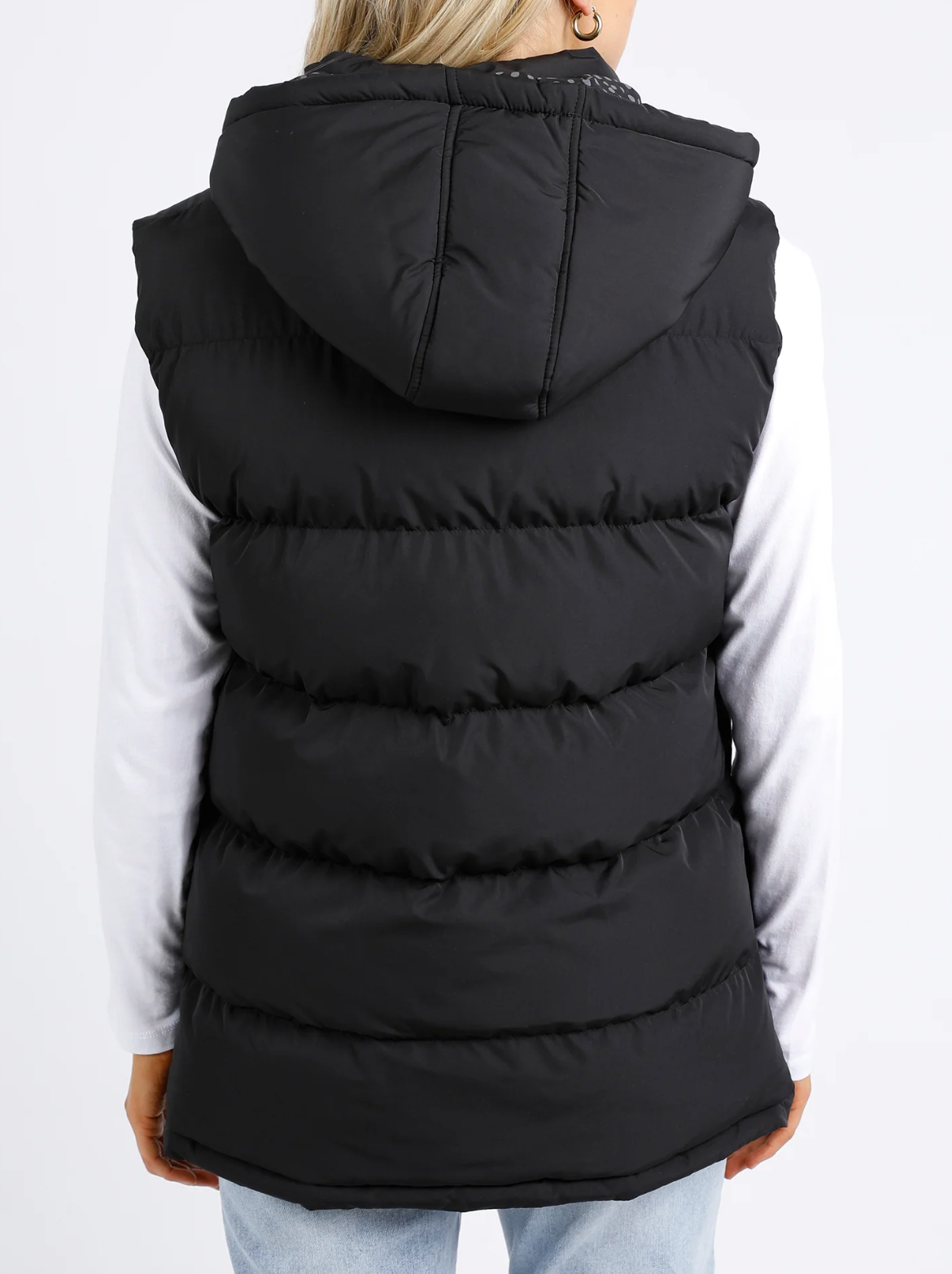 Rear view of a person in the Foxwood Camilla Puffer Vest, featuring quilted detailing and an adjustable hood, over a white long-sleeve shirt. The vest is paired with light blue jeans against a plain white background.