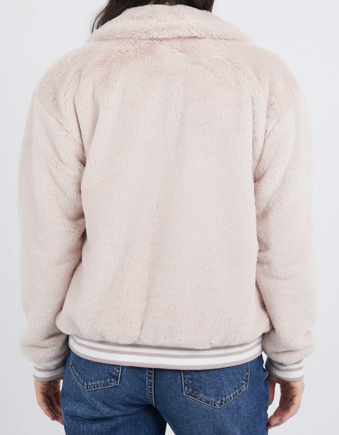 Wearing the Foxwood Nora Bomber, a person is seen from behind in a light pink, soft furry jacket made of 100% Polyester. It has long sleeves and a collar. They paired it with blue jeans against a plain white backdrop.