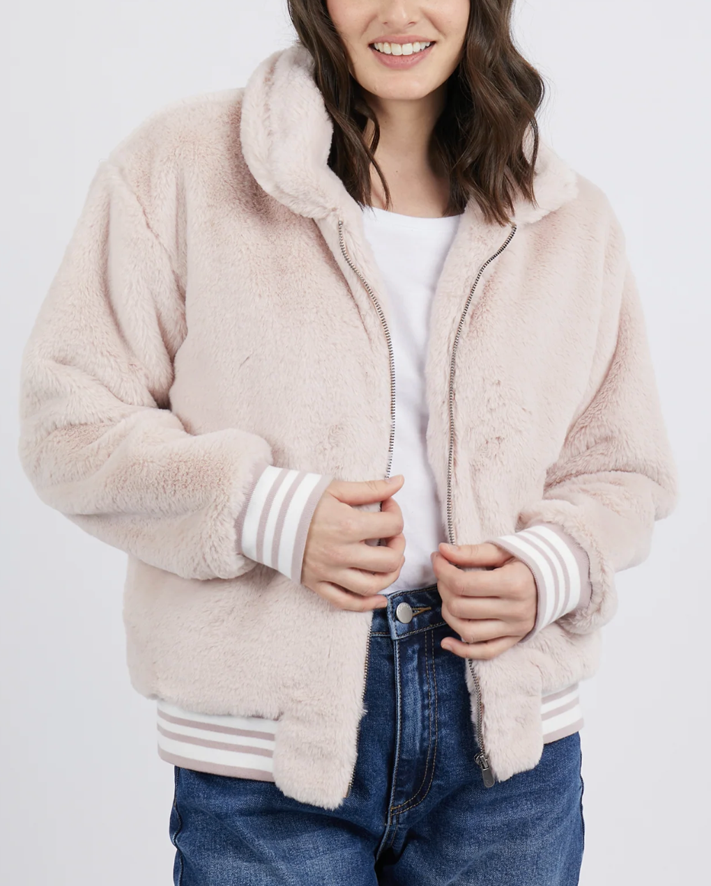 A person wears the Foxwood Nora Bomber, a light pink 100% polyester jacket with striped cuffs and a zipper, over a white shirt and blue jeans. The stylish piece complements their shoulder-length wavy hair as they smile with hands near the front zipper.