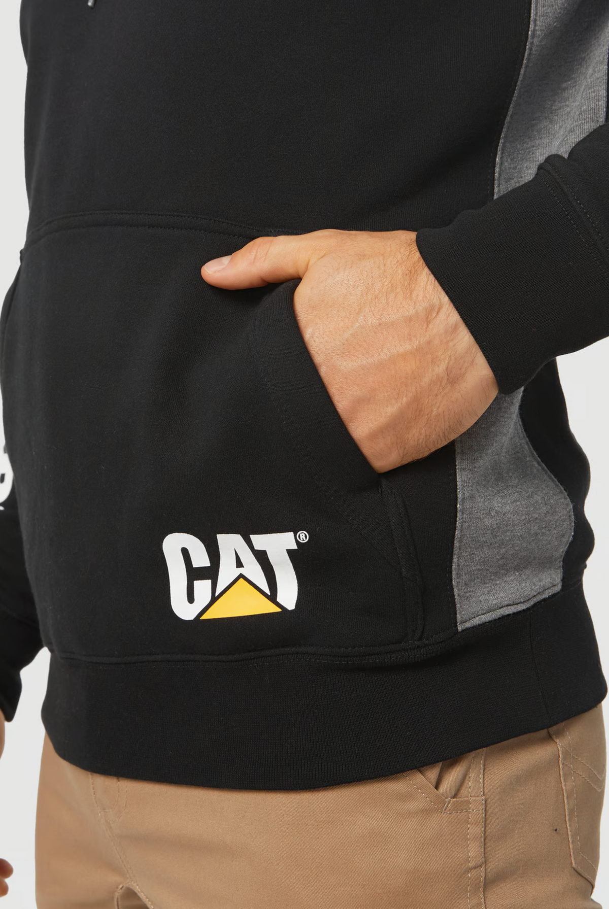 A person wearing a CAT Logo Panel Hooded Sweat in black and gray has the "CAT" logo on the front pocket where their right hand is tucked, paired with tan pants for a relaxed look.