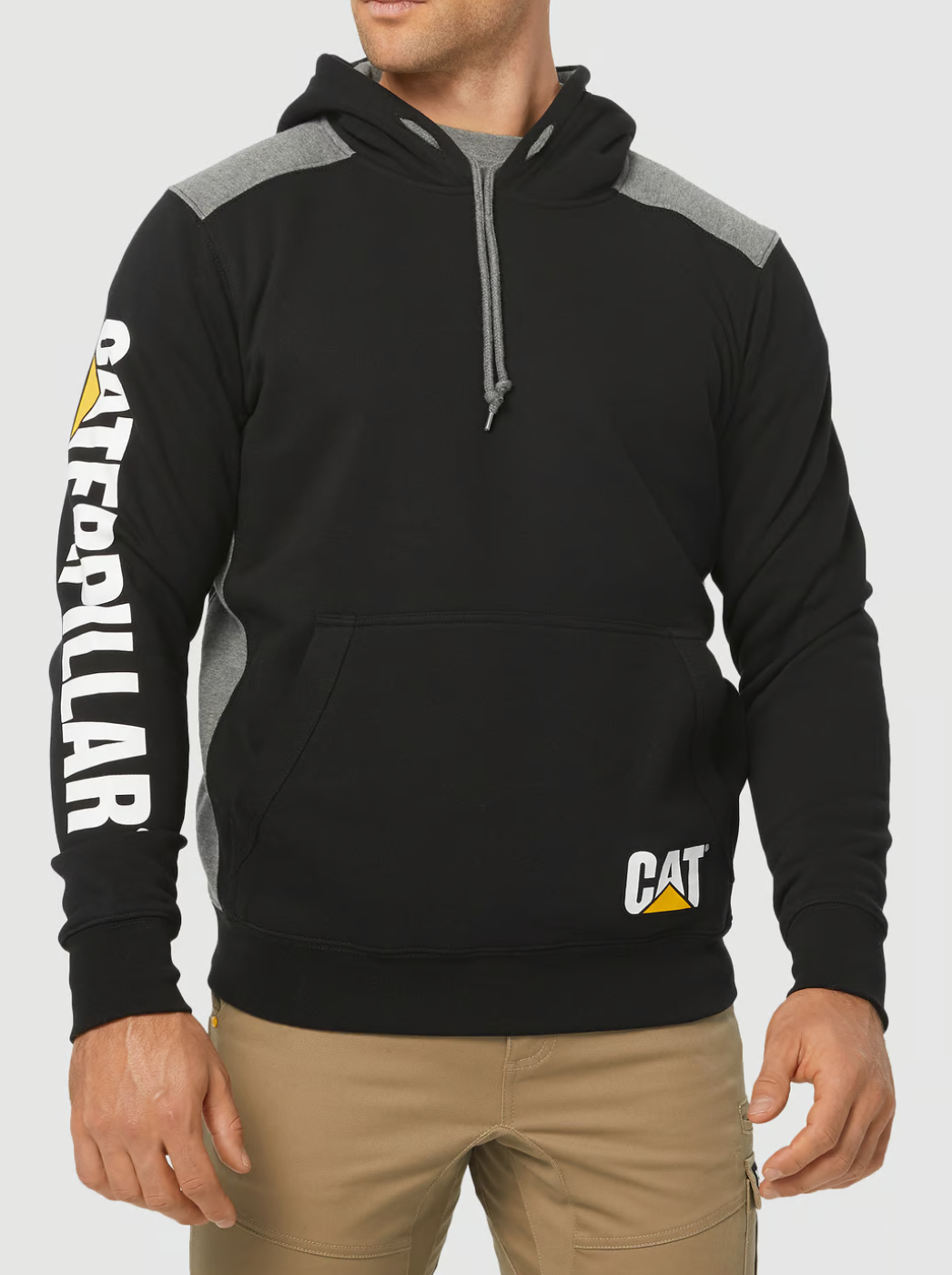On a plain white background, someone wears the CAT Logo Panel Hooded Sweat in black and gray with "CATERPILLAR" on the sleeve and the CAT logo on the front pocket. The hoodie, complete with a drawstring, is stylishly matched with beige pants.