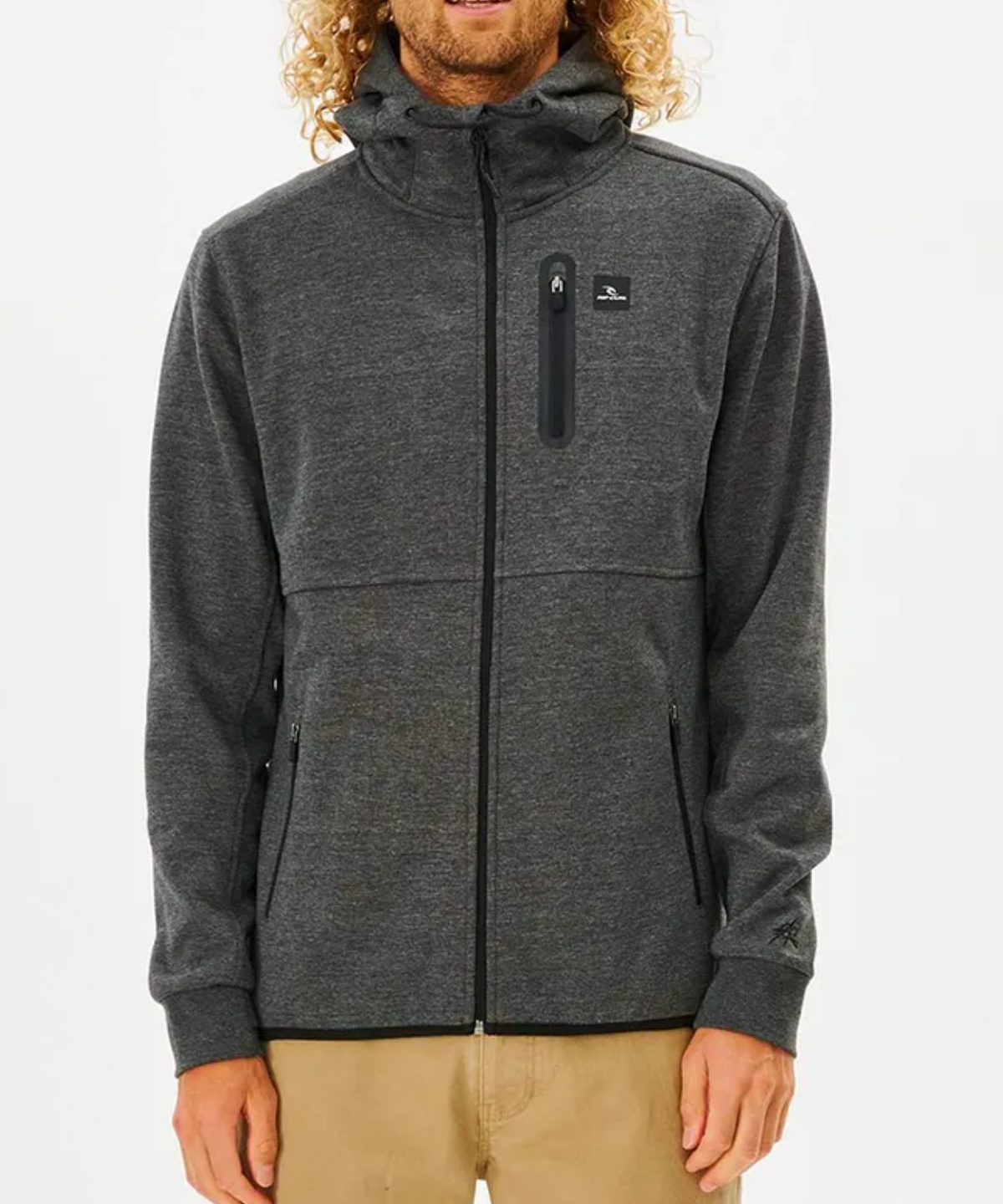 Against a white backdrop, someone wears the Rip Curl Anti Series Departed Zip Thru hoodie in dark gray, featuring a hood, zippered pockets, and chest logo. Paired with light brown pants, this stylish winter piece ensures warmth and fashion.