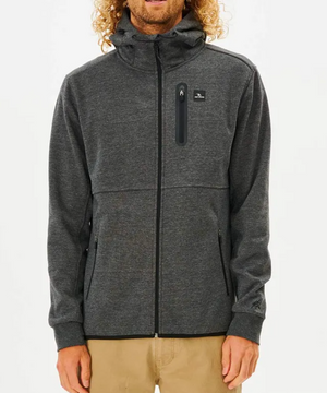 Against a white backdrop, someone wears the Rip Curl Anti Series Departed Zip Thru hoodie in dark gray, featuring a hood, zippered pockets, and chest logo. Paired with light brown pants, this stylish winter piece ensures warmth and fashion.