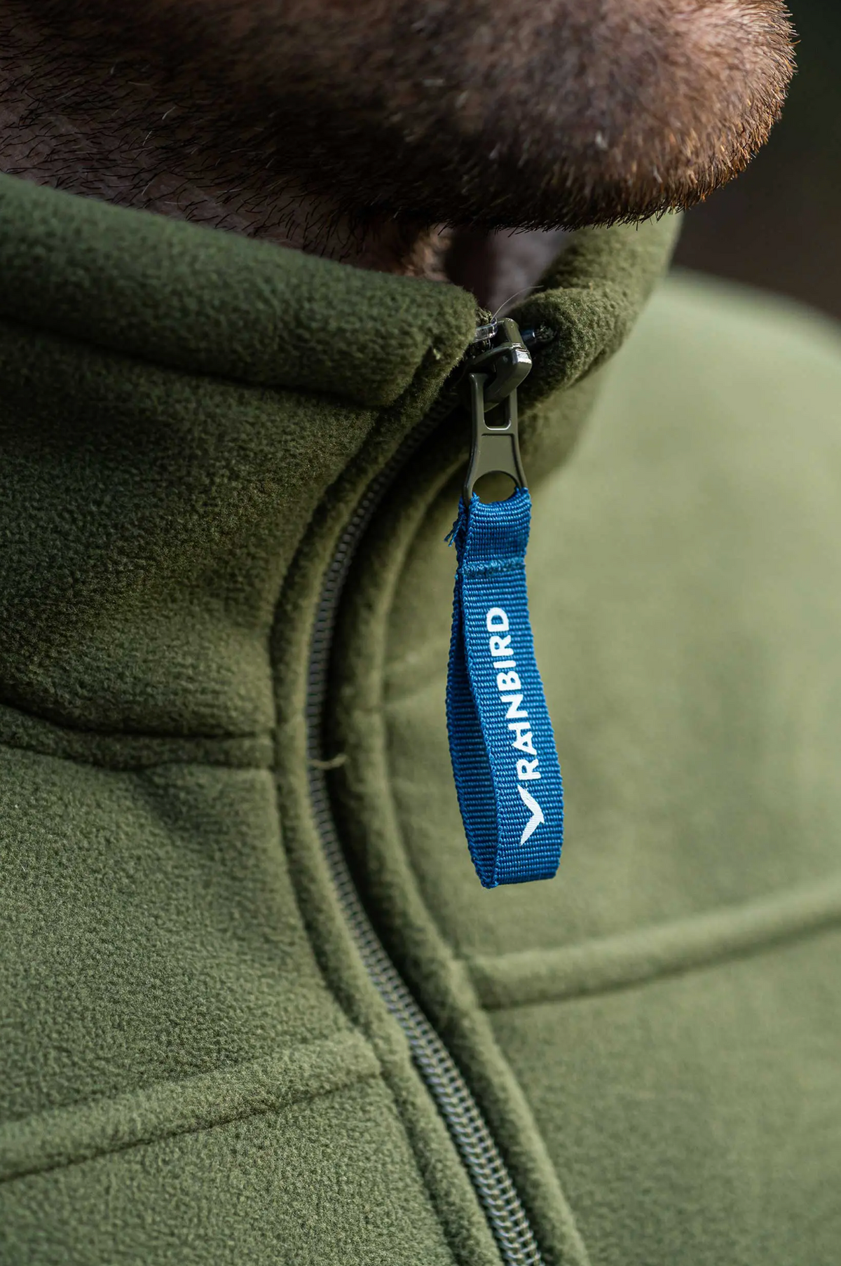 A close-up shows a person in a Rainbird Lumber Jacket, featuring a blue zipper pull marked "RAINBIRD." The focal point is the zipper area, with part of their chin seen above the collar. The warm polyester fleece lining ensures comfort in cooler weather.