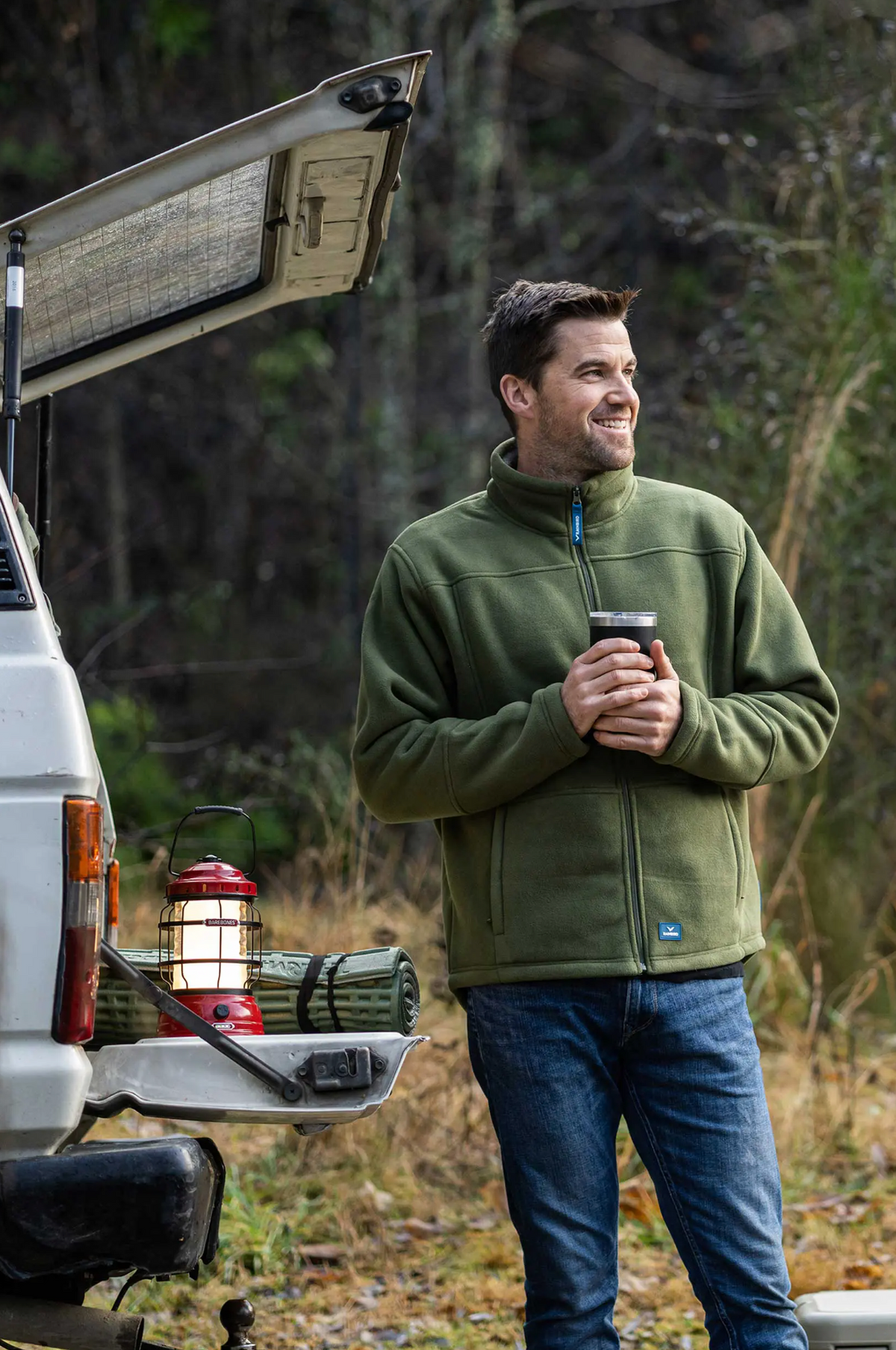 A man in a fleece-lined Rainbird Lumber Jacket leans against an open car trunk, holding a cup. A camping lantern and sleeping bag rest on the car's edge as he enjoys a leisurely moment outdoors in a wooded area.