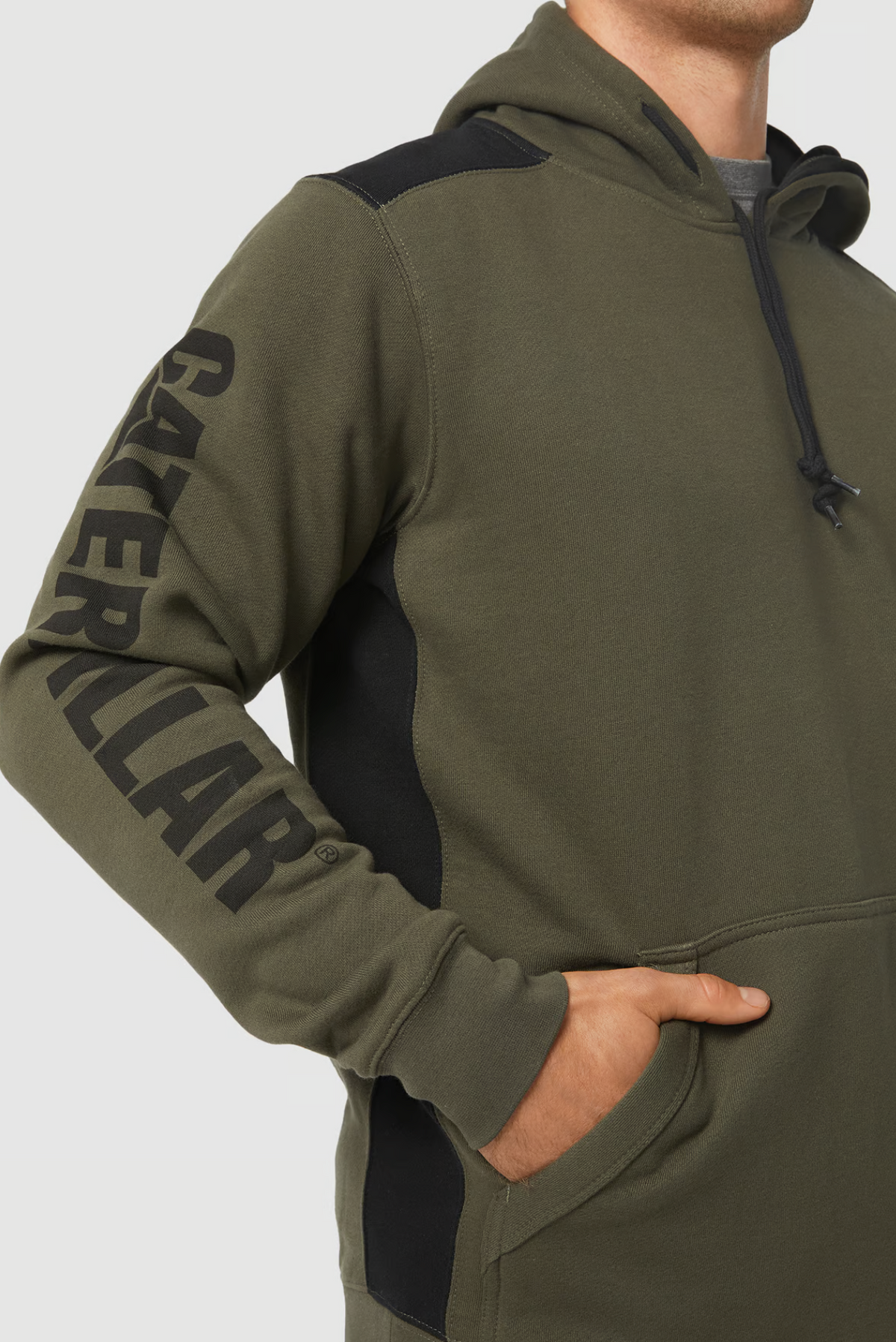 A person wearing a CAT Logo Panel Hooded Sweat in an olive green color with black accents on the shoulder and side. The left sleeve features "CATERPILLAR" printed in prominent black letters along its length. The person stands with a hand in the front pocket, which includes a hidden velcro phone pocket.