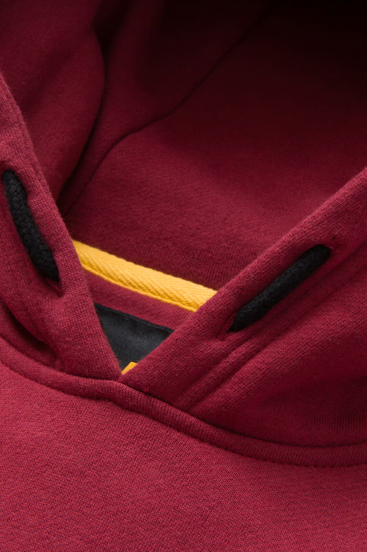 Close-up of the CAT Trademark Banner Hooded Sweat, a maroon hoodie with black drawstring holes and a subtle CAT logo. Yellow fleece lining peeks out beneath the rich maroon tone, offering a soft, cozy appeal.