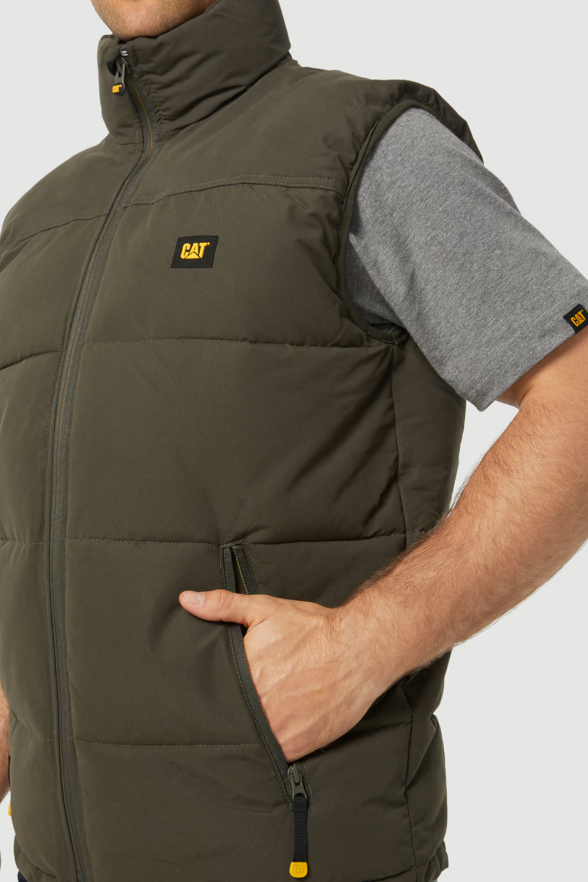 A person wearing a CAT Arctic Zone Vest in padded olive green with polyester insulation over a grey t-shirt, hand in the vest pocket, displaying the CAT logo on both the vest and sleeve.