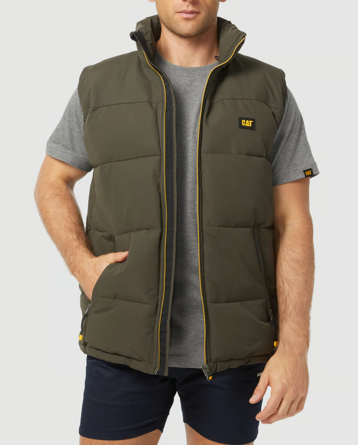 A man wears a gray t-shirt with dark shorts and the CAT Arctic Zone Vest, featuring a dark green color with yellow accents and "CAT" branding. The vest has an insulated stand-up collar and a water-resistant finish. His left hand rests in his front pocket against a plain white background.