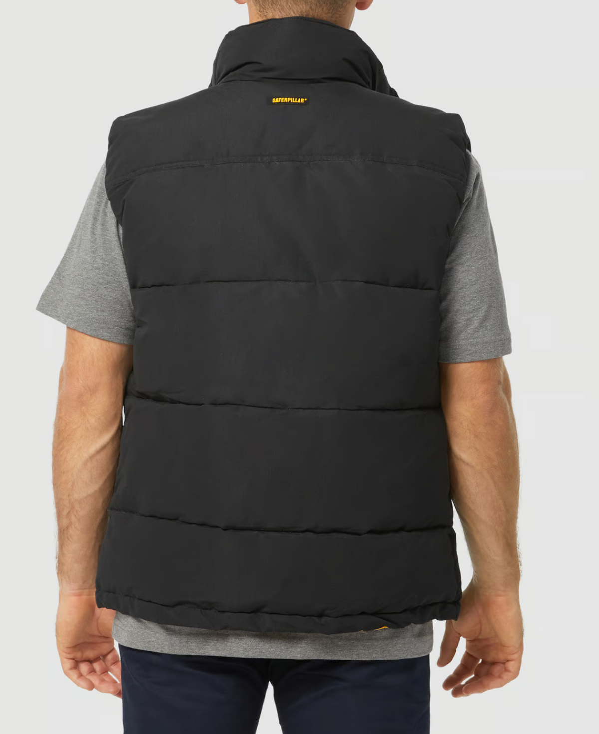 Rear view of a person wearing a black CAT Arctic Zone Vest, featuring a water-resistant finish, branded upper back, and insulated collar over a gray T-shirt. Paired with dark pants, they stand against a plain, light-colored background.