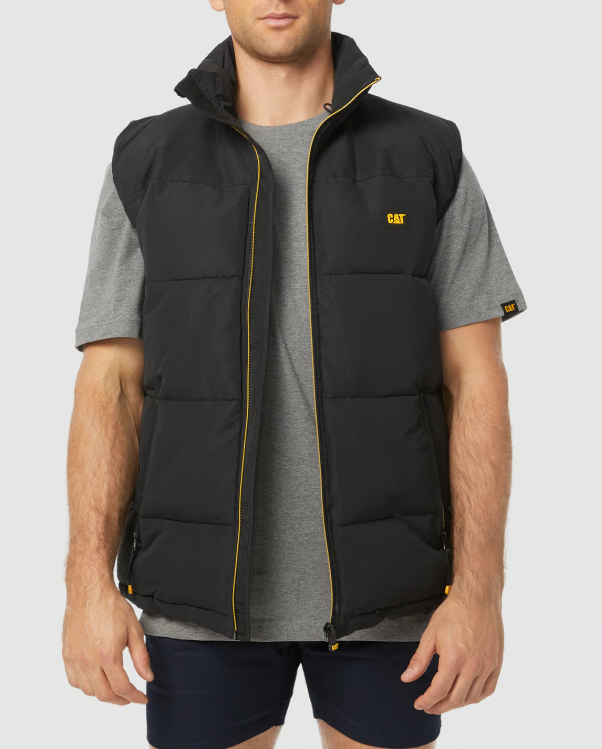 A man wearing an open CAT Arctic Zone Vest with yellow accents over a gray t-shirt and black shorts. The black sleeveless vest features an insulated collar, a water-resistant finish, and a small "CAT" logo on the front. The background is plain white.