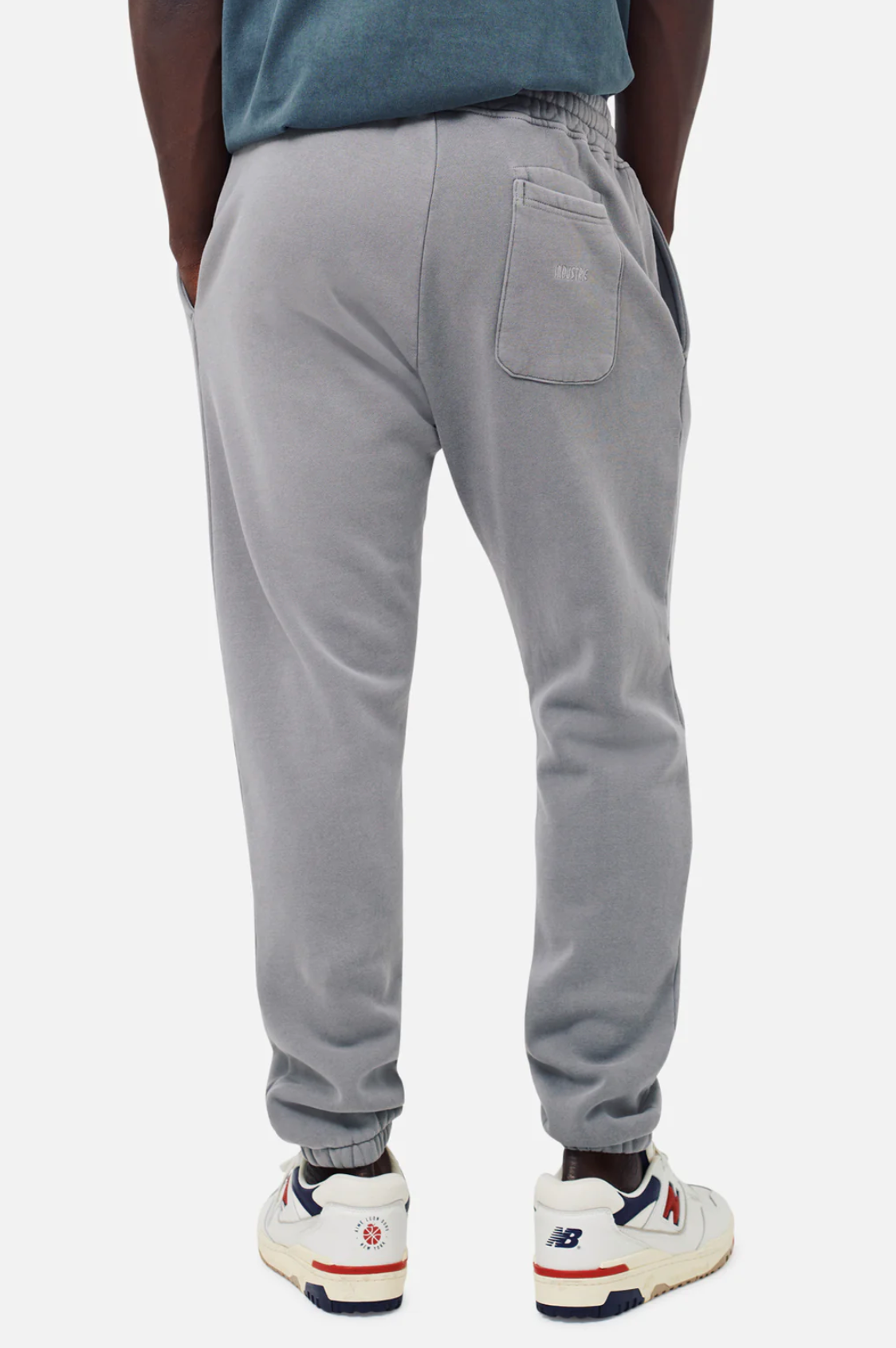 Person wearing Industrie The Del Sur Track Pant in light gray, hands in pockets, shown from behind. These joggers have an elastic waistband and one back pocket. They're paired with New Balance sneakers in white with red and navy accents, creating a casual style ideal for any outing.