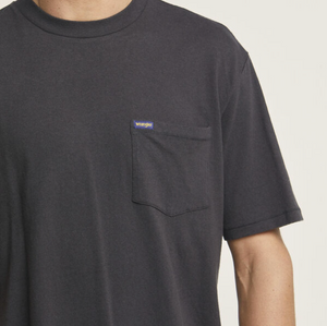 A man wears a Wrangler Classic Pocket Tee, in plain black with a small left-side pocket and tiny logo tag, made of 100% cotton. The simple setting highlights the casual tee on his upper body.