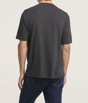 A person wearing a Wrangler Classic Pocket Tee in plain dark gray and blue jeans stands with their back turned. They have a small circular tattoo on their left arm, and the 100% cotton jeans have zigzag stitching on the pockets. The background is light-colored.