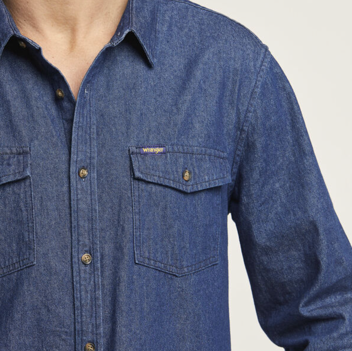 A person wears the Wrangler Roadside Denim Shirt, a regular fit blue denim made from 100% cotton. It features buttoned chest pockets, with a small logo on the left pocket against a plain, light background.