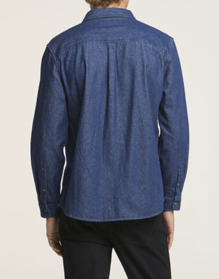 Rear view of a person in a Wrangler Roadside Denim Shirt, regular fit with long sleeves, paired with black pants. The blue denim shirt is 100% cotton, featuring a collar and buttoned cuffs against a plain white background.