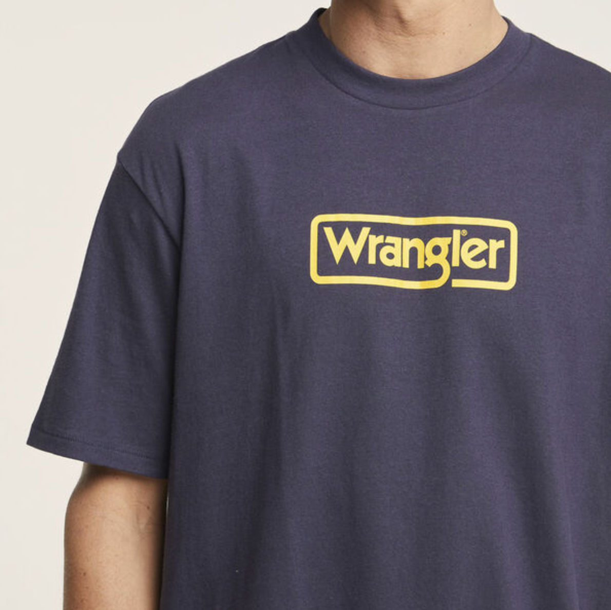 A person wears a Wrangler Border Tee, navy blue and 100% cotton, featuring a yellow logo on the chest. The light-colored background is plain, and the person's face is not visible.