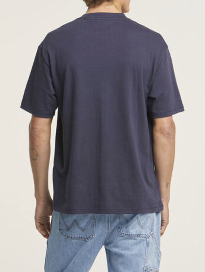 A person is standing with their back to the camera, wearing a dark blue Wrangler Border Tee and light blue jeans with distinctive stitching on the back pockets. The 100% cotton shirt complements the jeans, and the regular fit adds comfort against a plain, light-colored background.