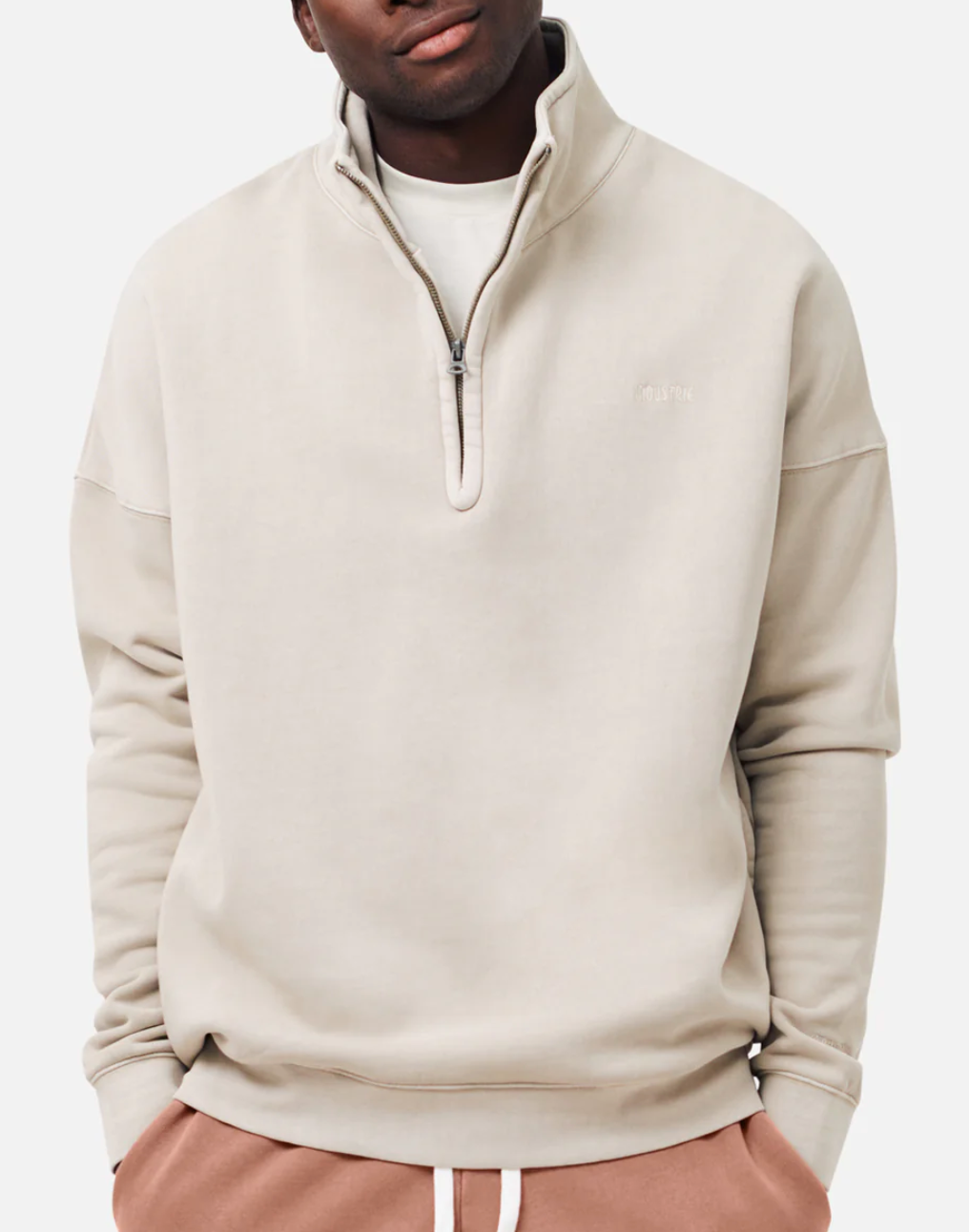 A person in an oversized, light beige Industrie The Washed Del Sur Track Top and peach-colored pants stands against a plain background. Their hands are in their pockets, with a white shirt peeking at the collar under the track top.