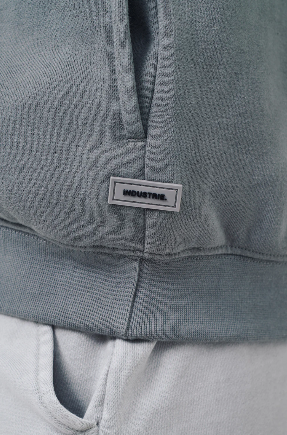 Close-up of a person wearing the Industrie The Del Sur Track Top, an oversized muted green hoodie with soft fleece lining. It features a small rectangular "INDUSTRIE" label and ribbed cuffs, paired with light gray pants.