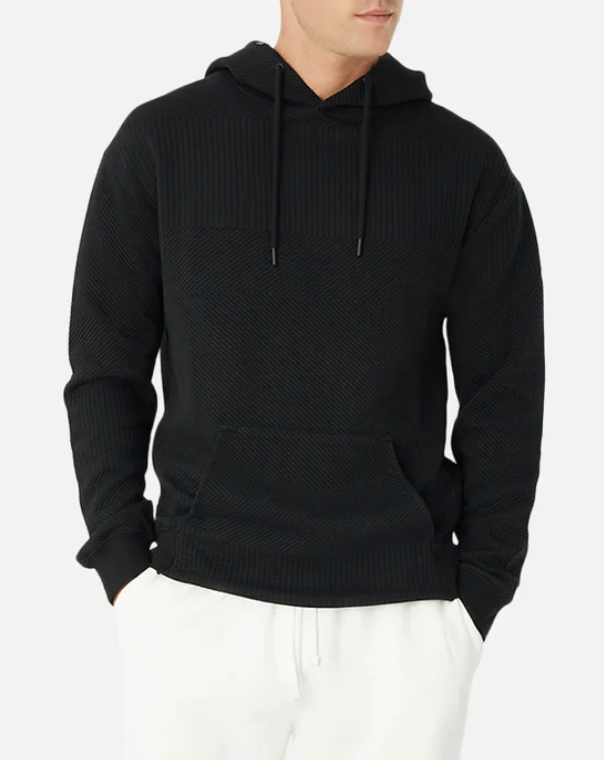 A person in an Industrie The Robinson Hoodie, featuring a black, relaxed fit with a textured design and drawstrings, paired with white pants. Hands rest in the hoodie's front pocket against a light-colored background.
