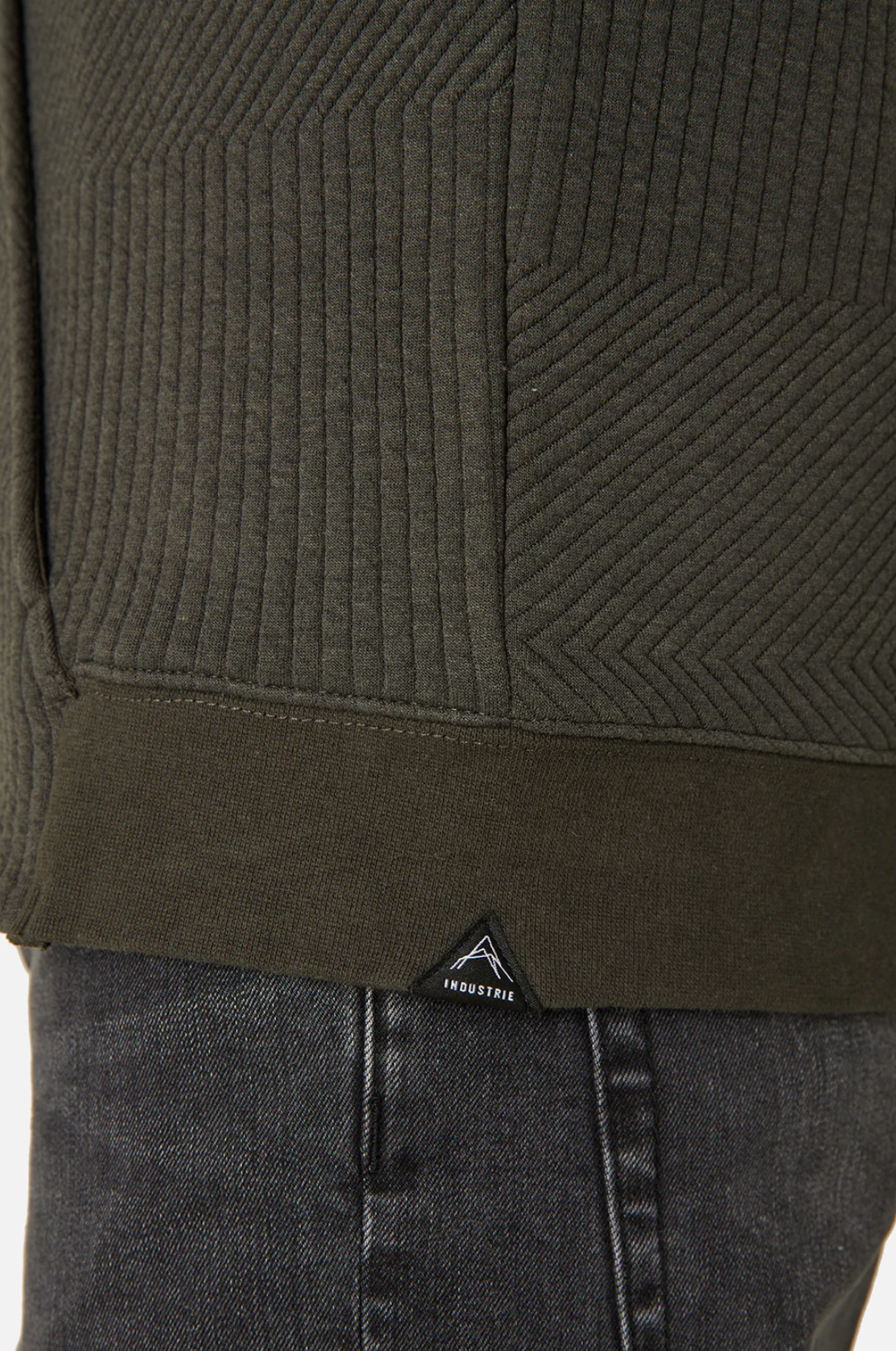 A close-up shows a person in Industrie The Robinson Hoodie, a textured dark green jacket with diagonal patterns and a relaxed fit. A small triangular INDUSTRIE logo is near the hem, and the bottom overlaps dark denim jeans for casual sophistication.