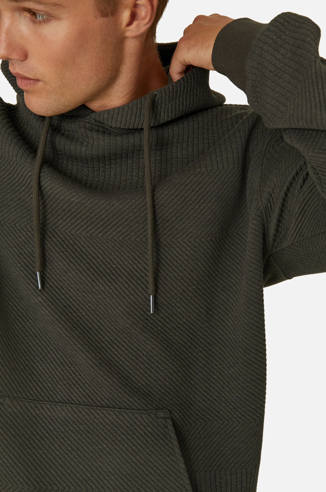 Wearing the Industrie The Robinson Hoodie, a person adjusts the hood of their dark ribbed hoodie with a textured design. It features a relaxed fit, drawstrings, and a kangaroo pocket against a plain white background with their face partially visible.