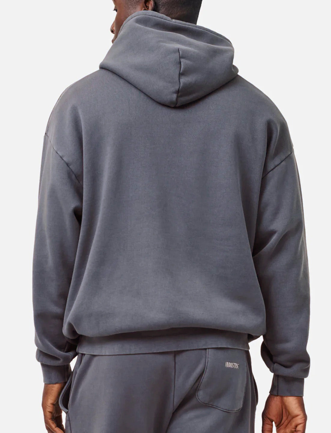 A person is seen from behind wearing the Industrie The Del Sur Hoodie in acid wash gray, paired with matching sweatpants. The loose-fitting hoodie features a hood, while the sweatpants have an embroidered text on the back pocket. They stand against a plain light gray background.